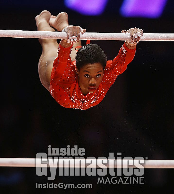 An Inside Gymnastics photograph of Gabby Douglas swinging through a handstand on the uneven bars in a red leotard, looking straight down the lens.
