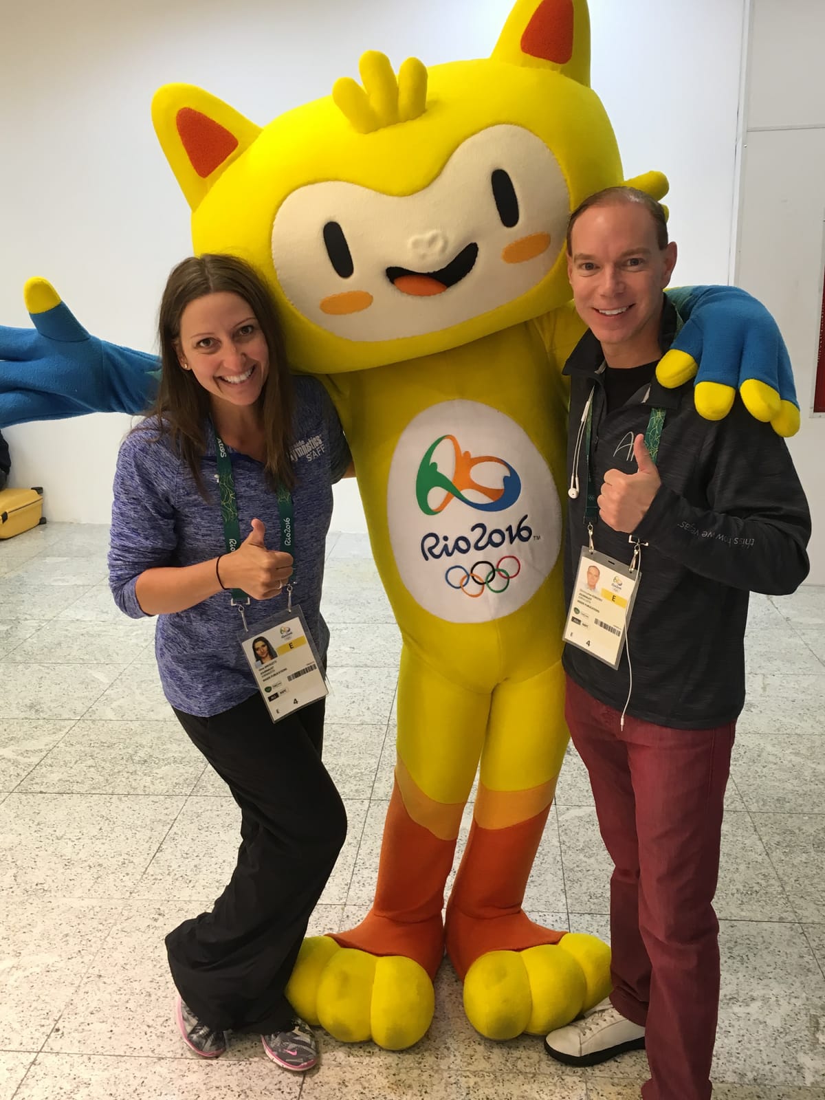Gina Pongetti Angeletti and a colleague giving thumbs up on either side of Vinicius, the yellow Rio 2016 mascot, both wearing Games credentials.