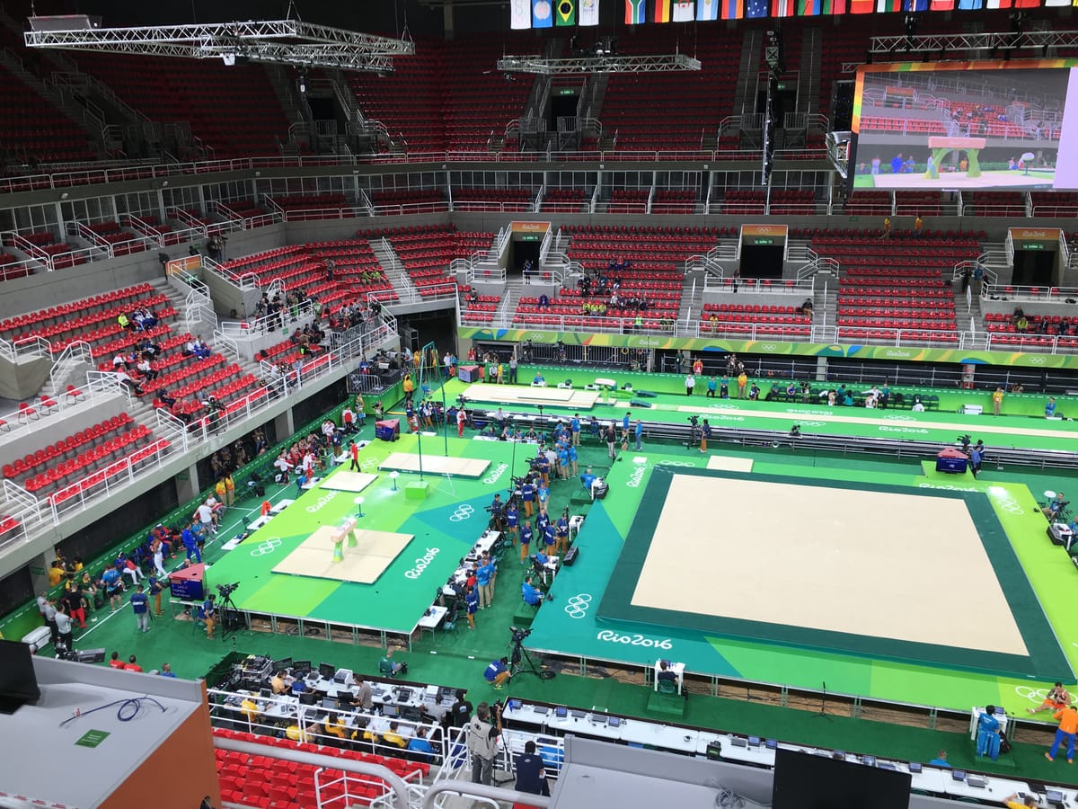 The Rio gymnastics arena seen from the stands during podium training: green and cream floor, bars and vault runway, camera crews at the edge, red seats mostly empty.