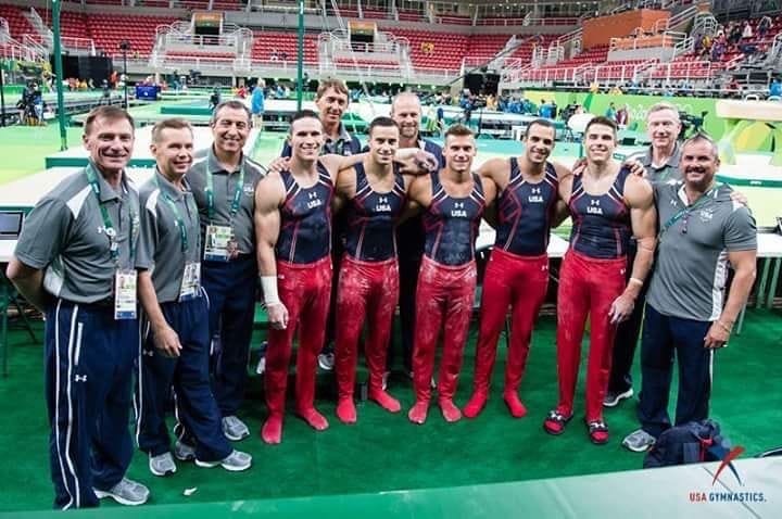 The five US men's gymnasts in Team USA competition uniforms lined up on the green podium with their coaching and medical staff.