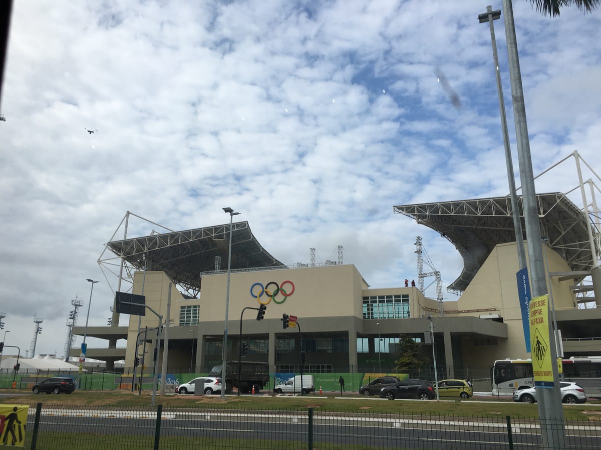 The exterior of the Rio Olympic arena with the coloured Olympic rings on its facade, traffic and a bus passing on the road in front.