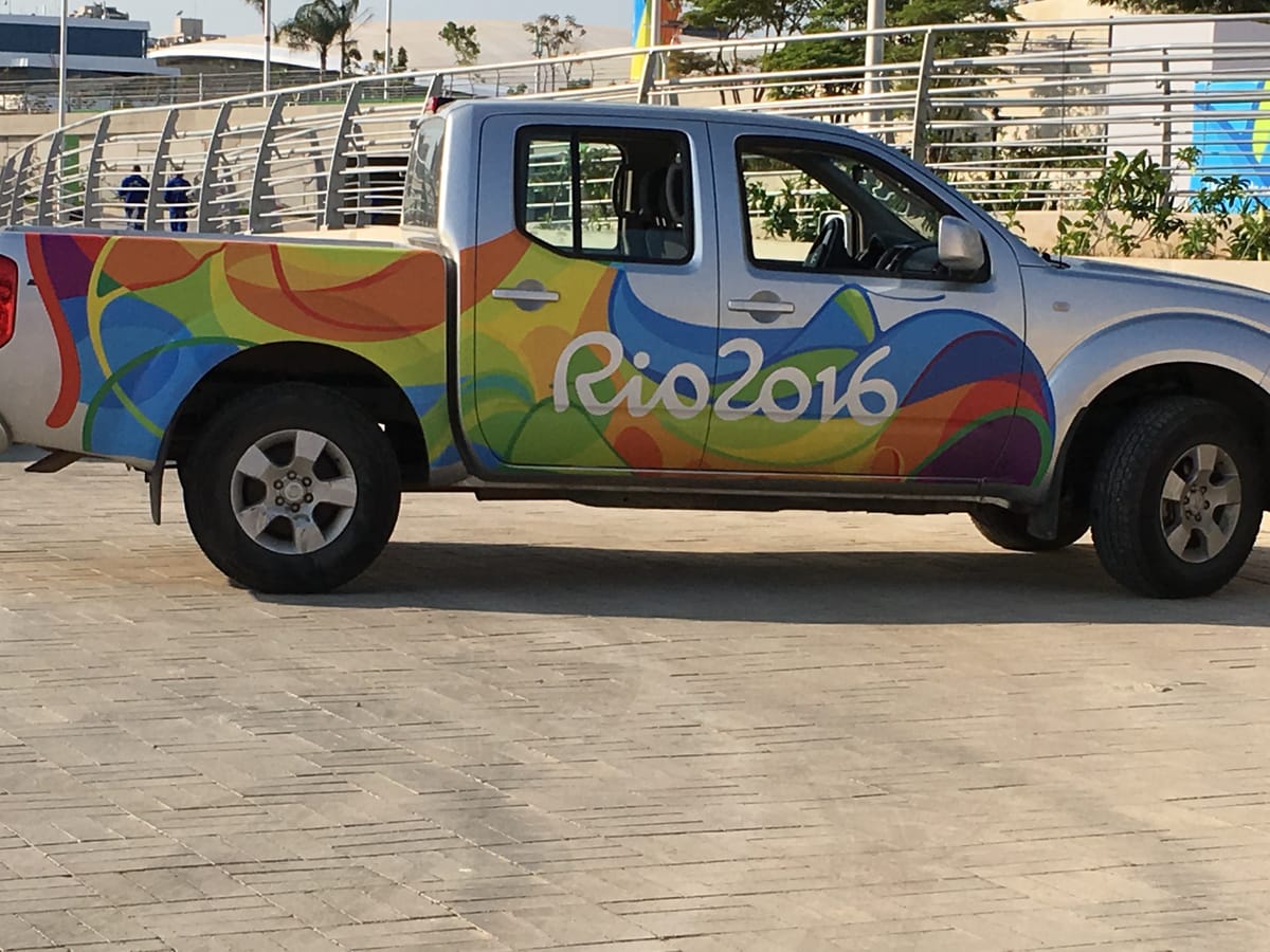 A silver pickup truck wrapped head to tail in the multicoloured Rio 2016 livery, parked on a paved plaza in the Olympic Park.
