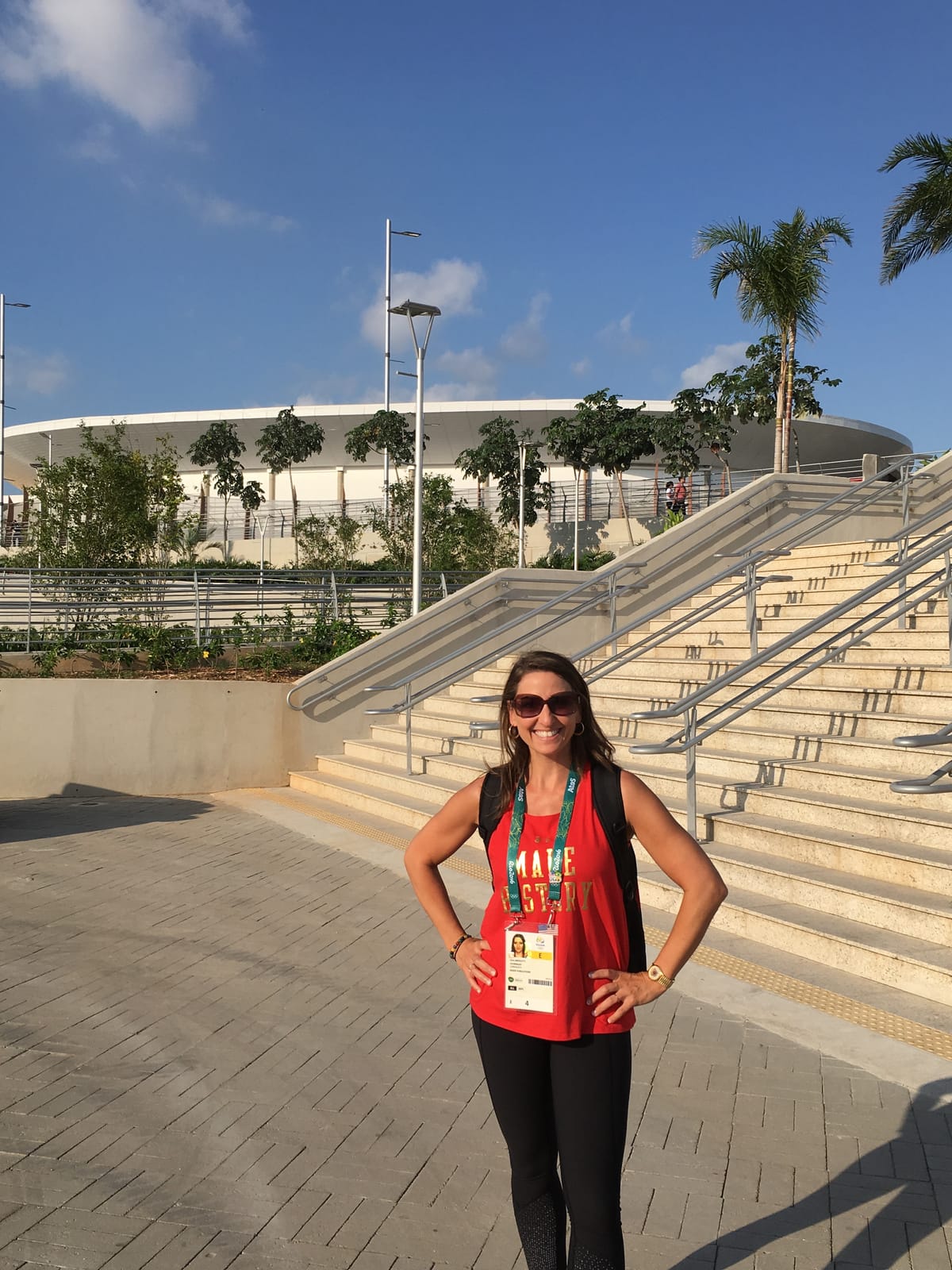 Gina Pongetti Angeletti in a red tank top and Rio credential at the foot of a wide staircase outside a Rio venue, palm trees and the arena roof behind her.