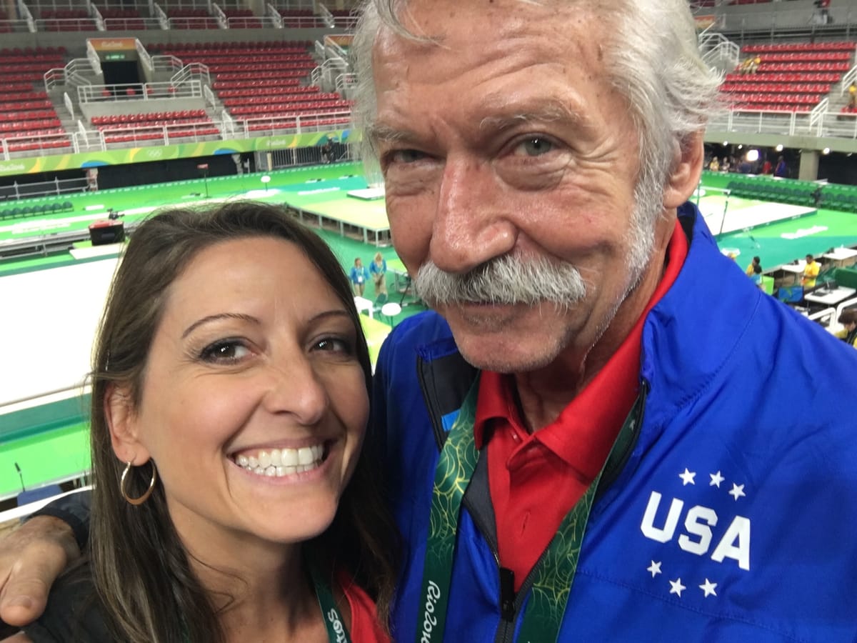 Gina Pongetti Angeletti cheek to cheek with Bela Karolyi in a blue USA jacket, the green competition floor of the Rio arena behind them.