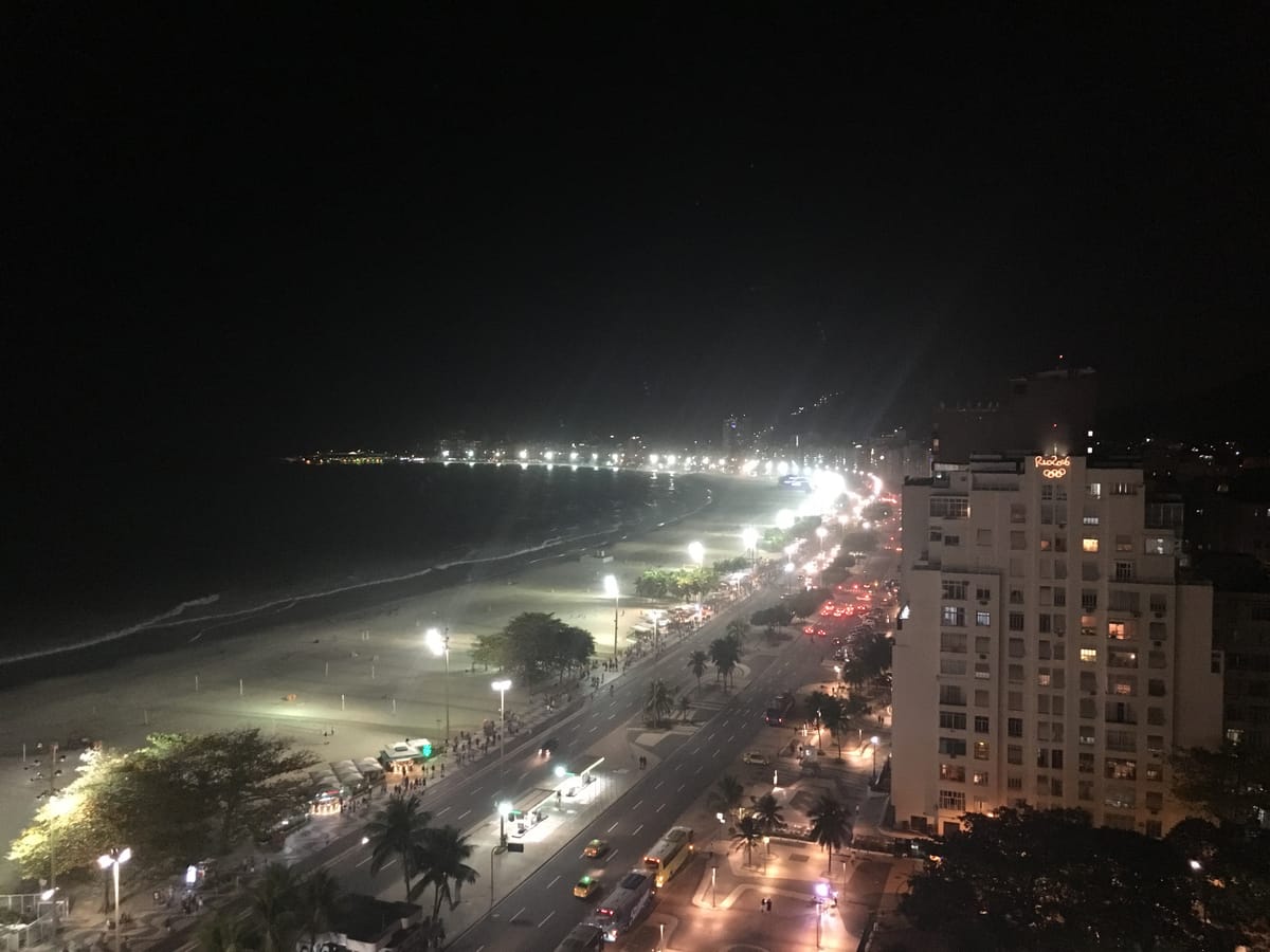 Copacabana at night from above: the lit promenade and beach on one side, traffic and apartment blocks on the other, one rooftop carrying an illuminated Rio 2016 sign.