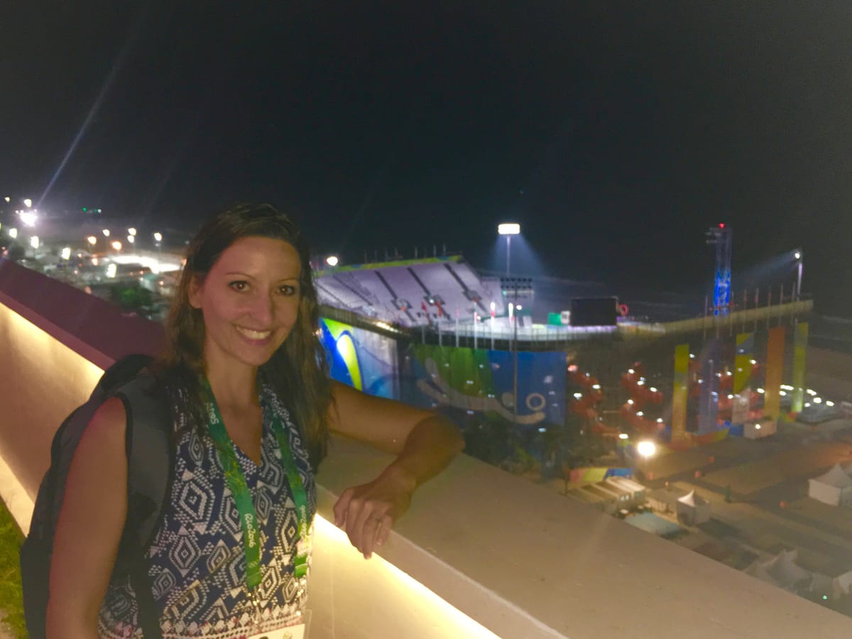 Gina Pongetti Angeletti on a balcony at night with the floodlit Olympic beach volleyball stadium on Copacabana behind her.