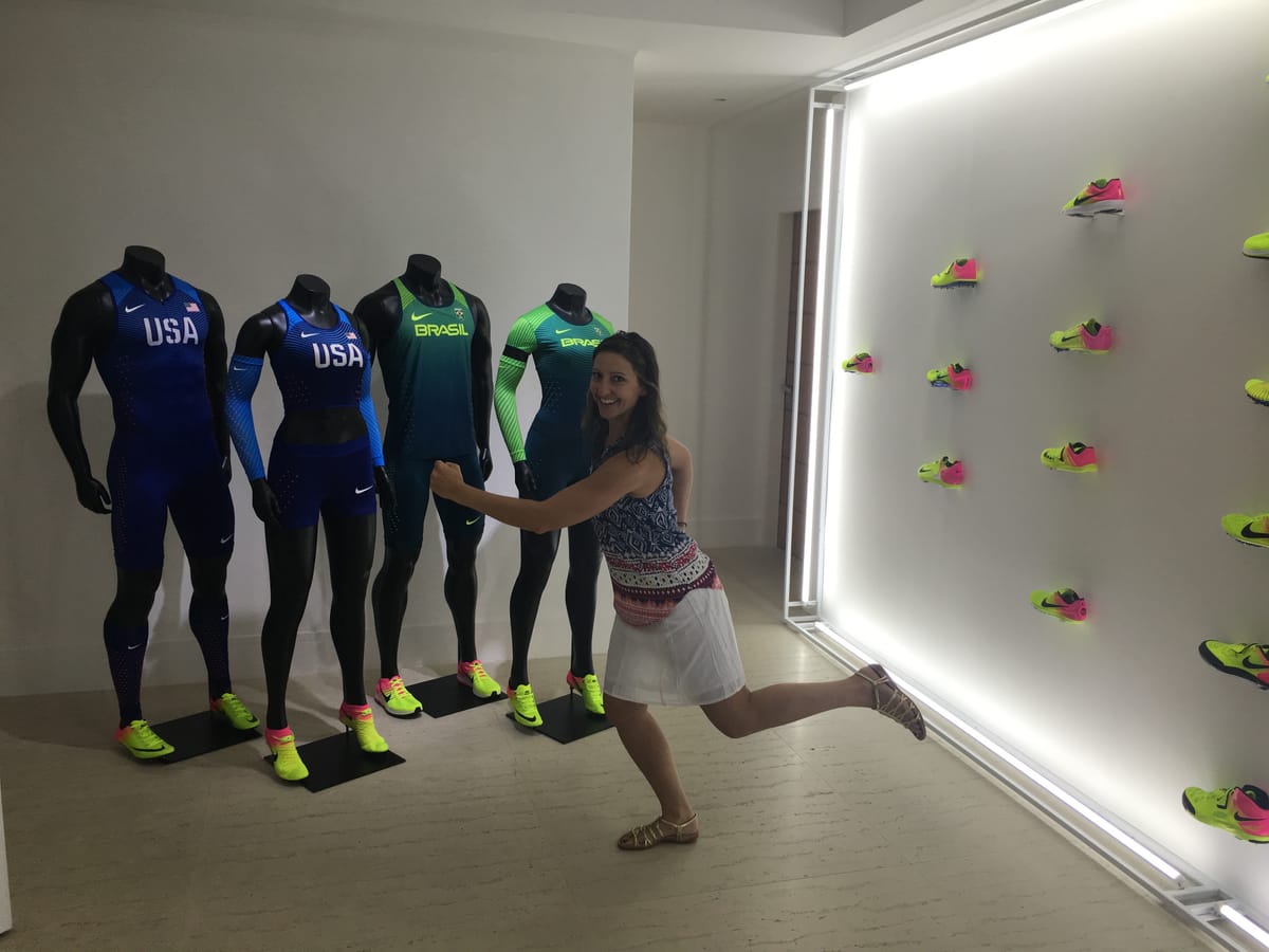 Gina Pongetti Angeletti striking a sprinter's pose beside four mannequins in Nike USA and Brasil track kit, a wall of neon running spikes alongside.