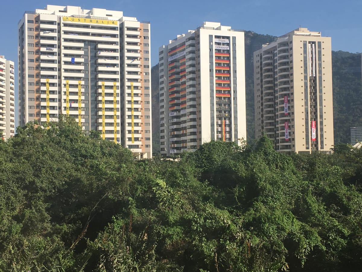 Three athlete village tower blocks rising above dense green trees, hung with national banners for Australia and Team Germany.