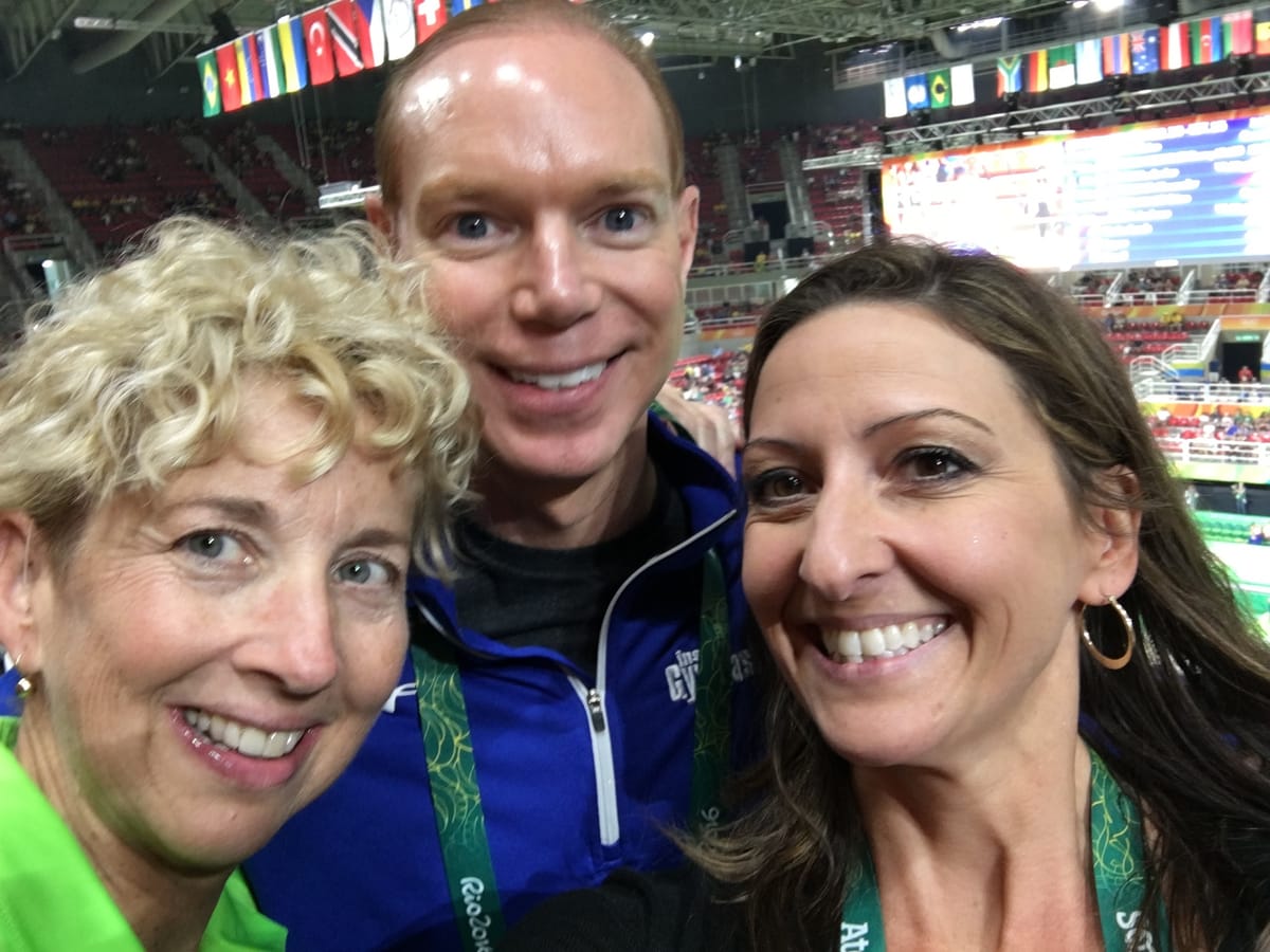 Gina Pongetti Angeletti with two Inside Gymnastics colleagues in the stands, national flags strung across the arena roof behind.