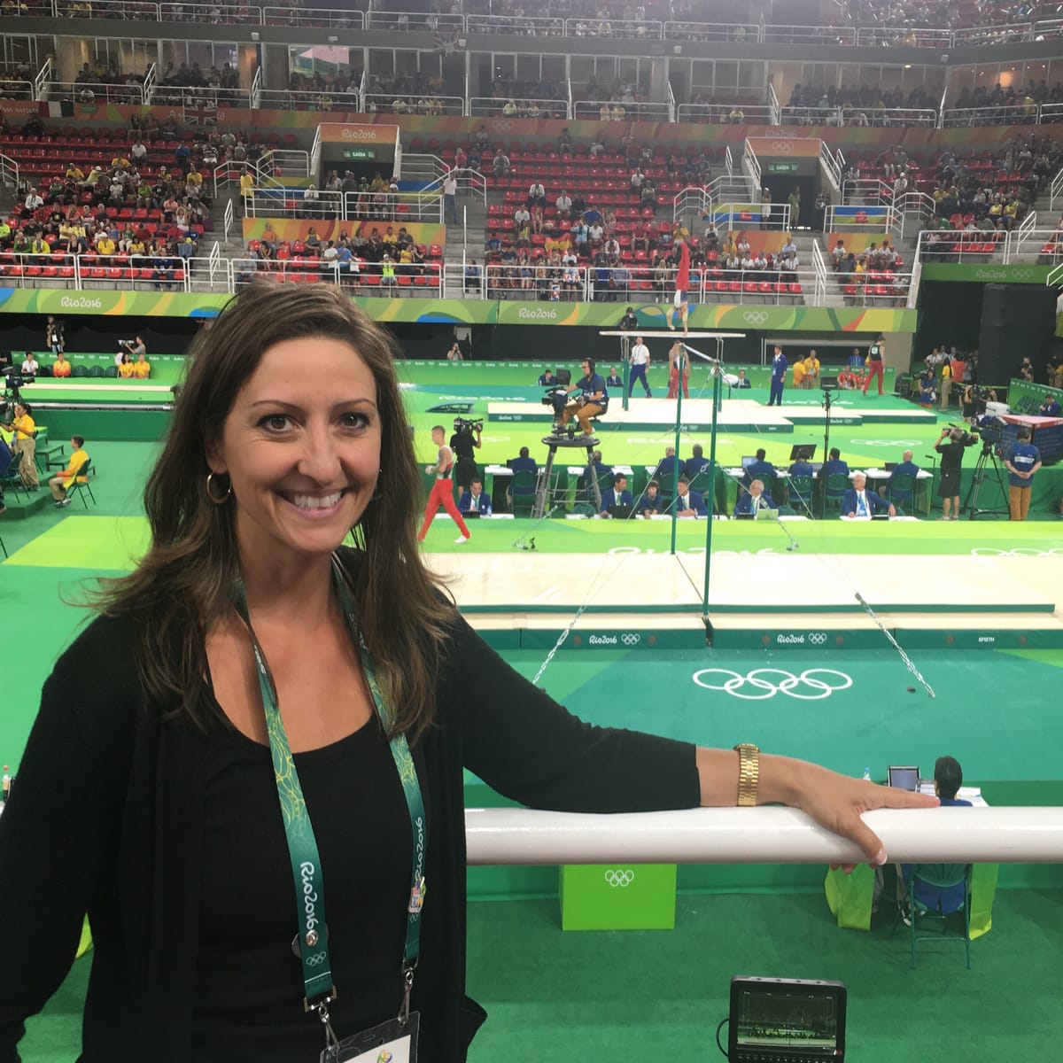 Gina Pongetti Angeletti at the press rail inside the Rio arena, the uneven bars, the judges' tables and a gymnast mid-routine behind her.
