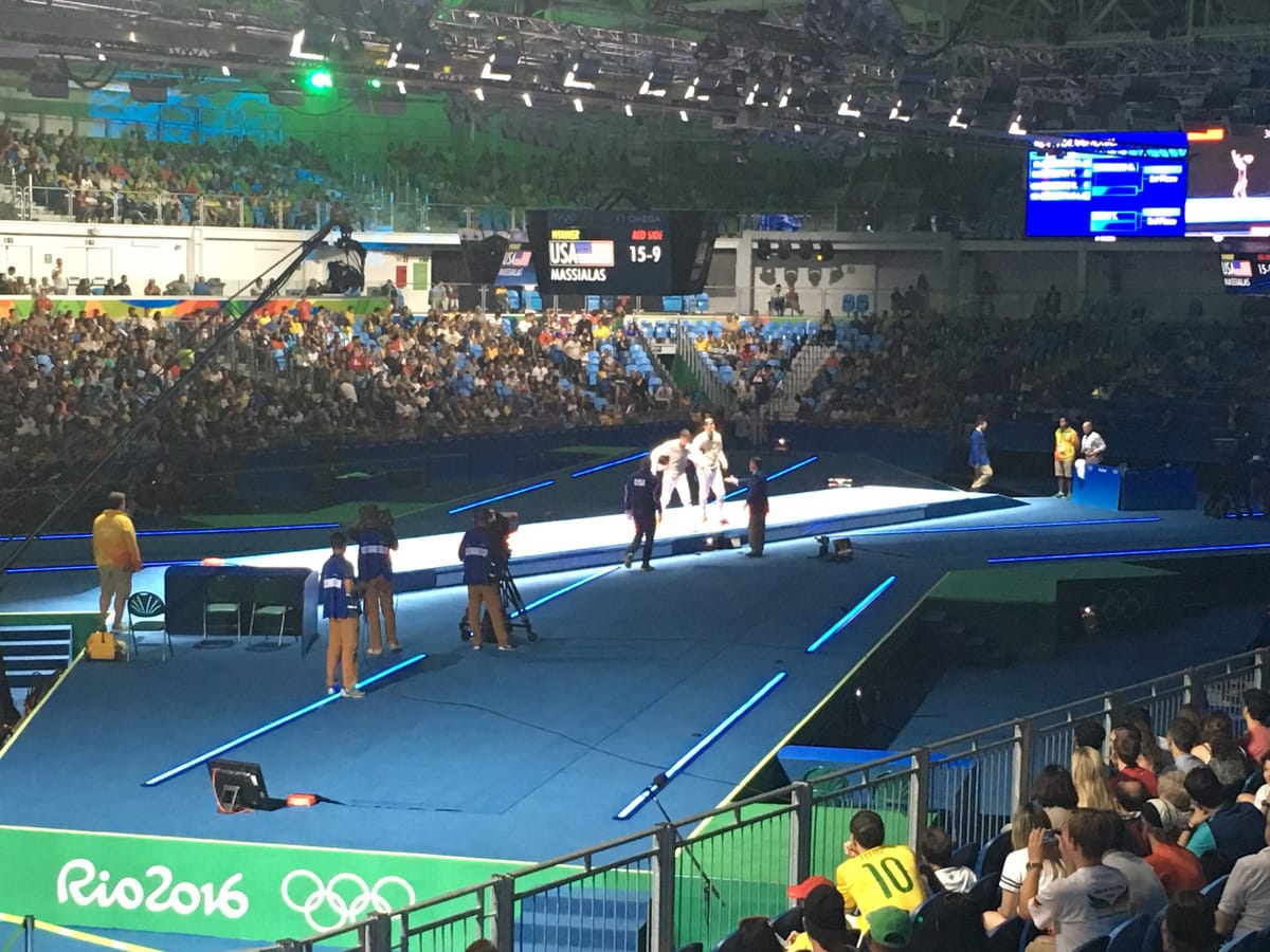 Olympic fencing: two fencers on the lit piste with the scoreboard reading USA MASSIALAS 15-9, spectators packed around the arena.