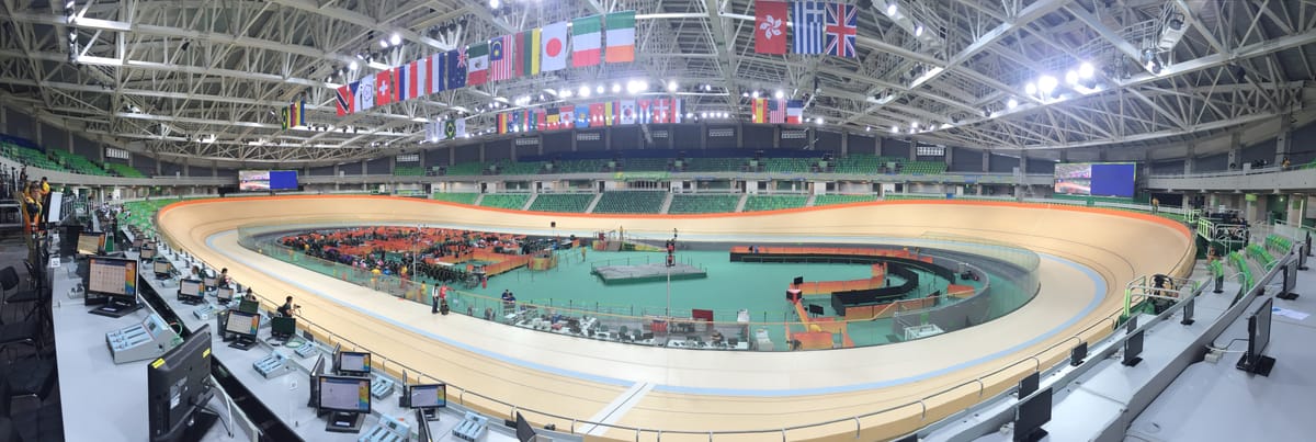 A panorama inside the Rio velodrome, the empty wooden track curving away, national flags overhead and commentary positions in the foreground.