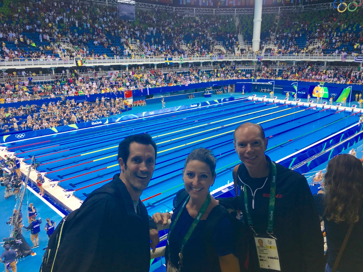 Gina Pongetti Angeletti and two colleagues in the stands at the Olympic Aquatics Stadium, the blue pool and a packed crowd behind them.