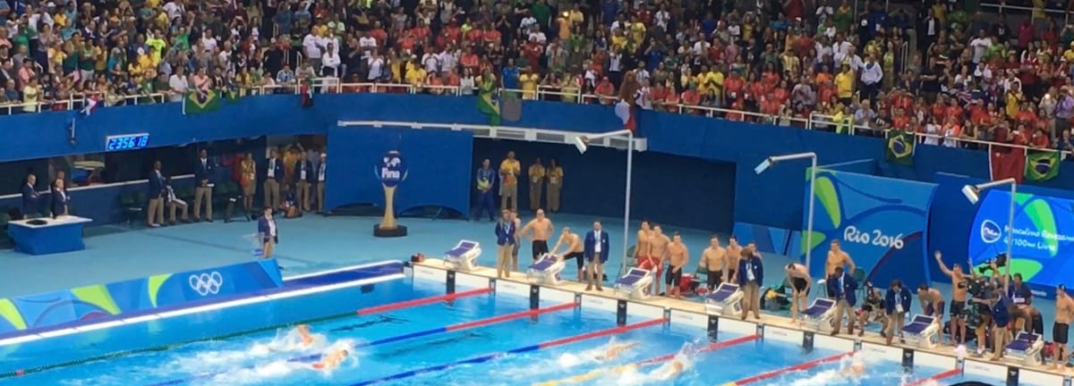 A swimming heat under way in the Olympic pool, swimmers mid-race and teams standing behind the blocks.