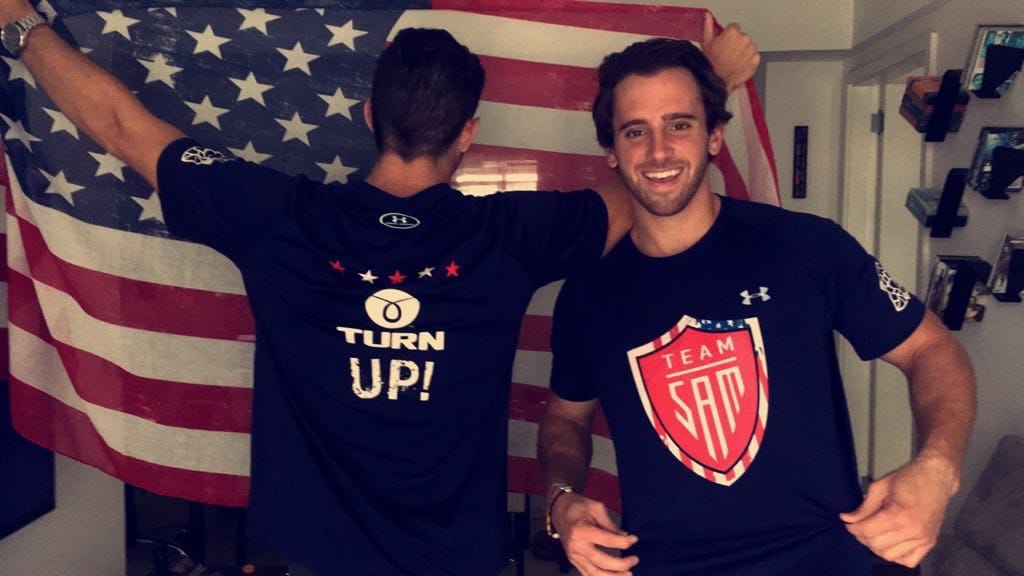 Two young men in front of a US flag hung on a wall, one turned away in a TURN UP! shirt, the other facing the camera in a TEAM SAM shirt.