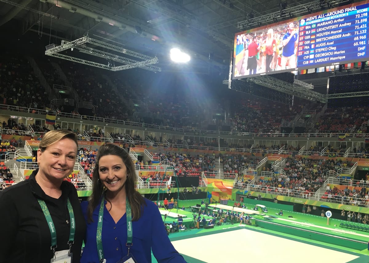 Gina Pongetti Angeletti and Peggy Liddick in the arena stands during the men's all-around final, the big screen behind them showing the standings after rotation five.