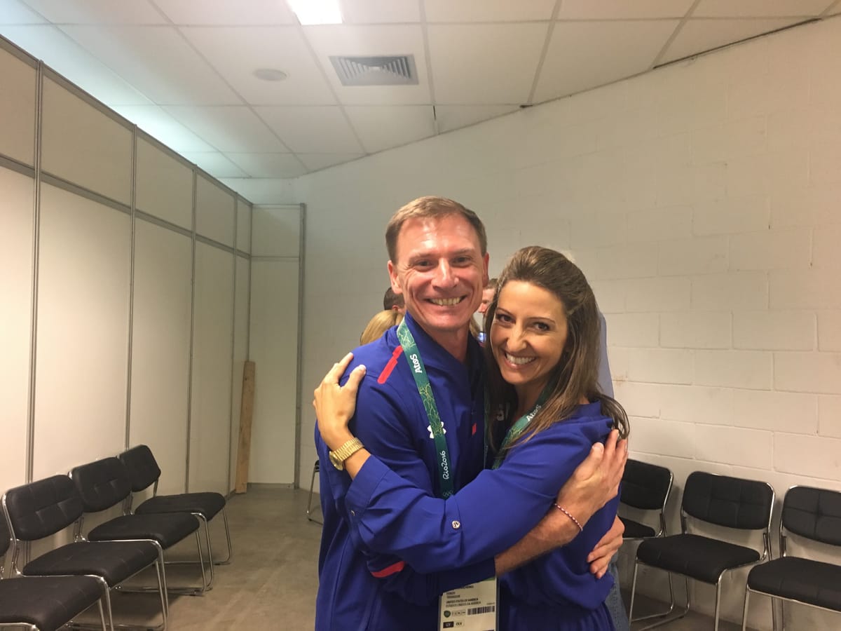 Gina Pongetti Angeletti and a man in a blue Team USA jacket hugging and laughing in a bare backstage room lined with stacking chairs.