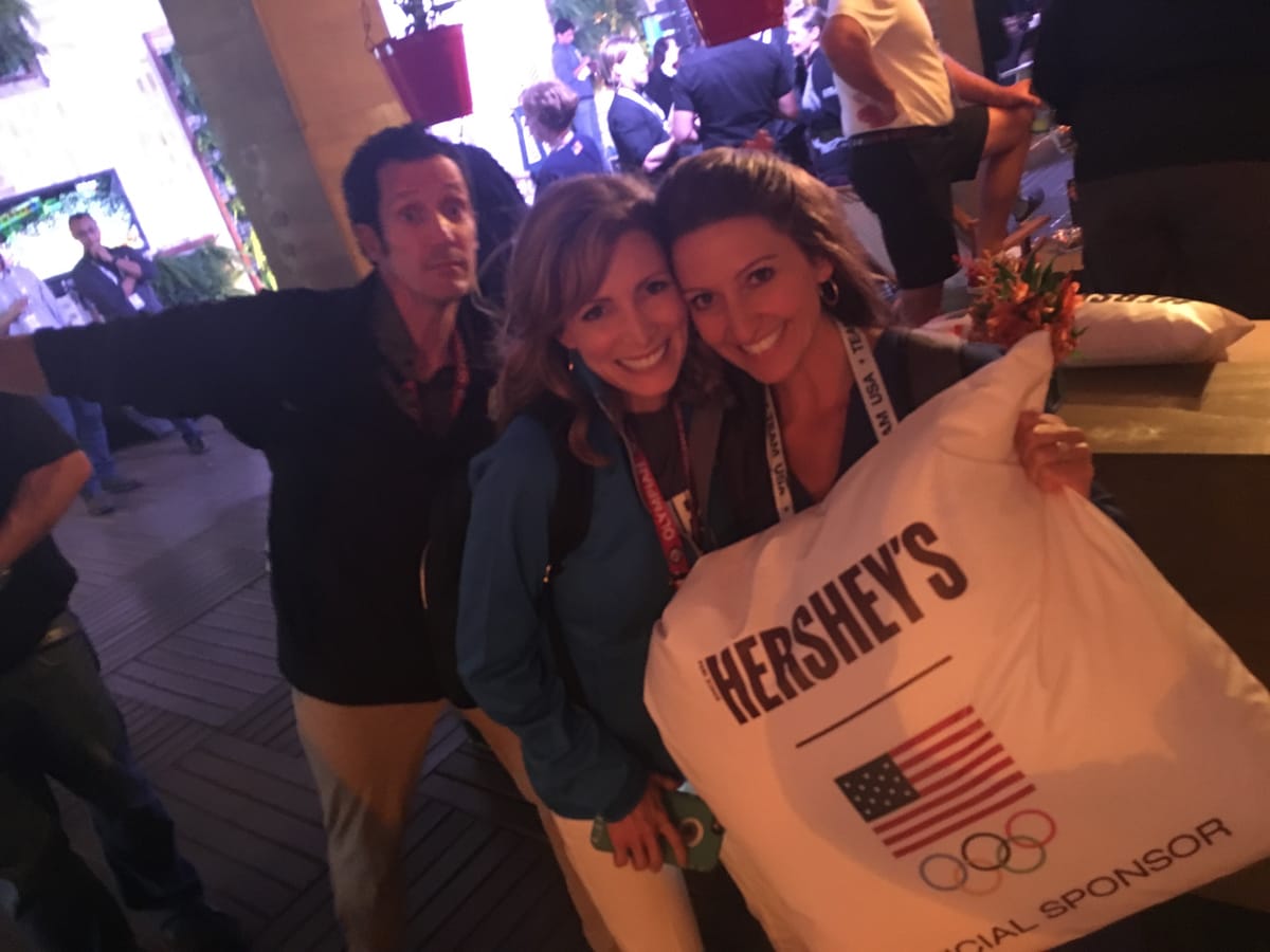 Gina Pongetti Angeletti and a friend cheek to cheek at a Team USA event holding a white Hershey's official sponsor cushion, a third person photobombing behind.
