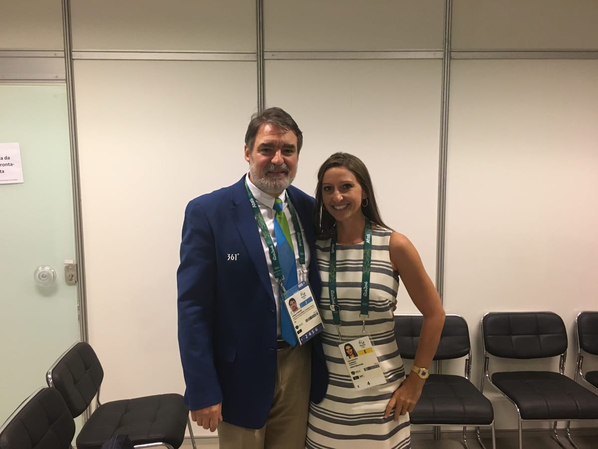 Gina Pongetti Angeletti with a bearded man in a blue Rio 2016 blazer and Games credential in a bare back-of-house room.