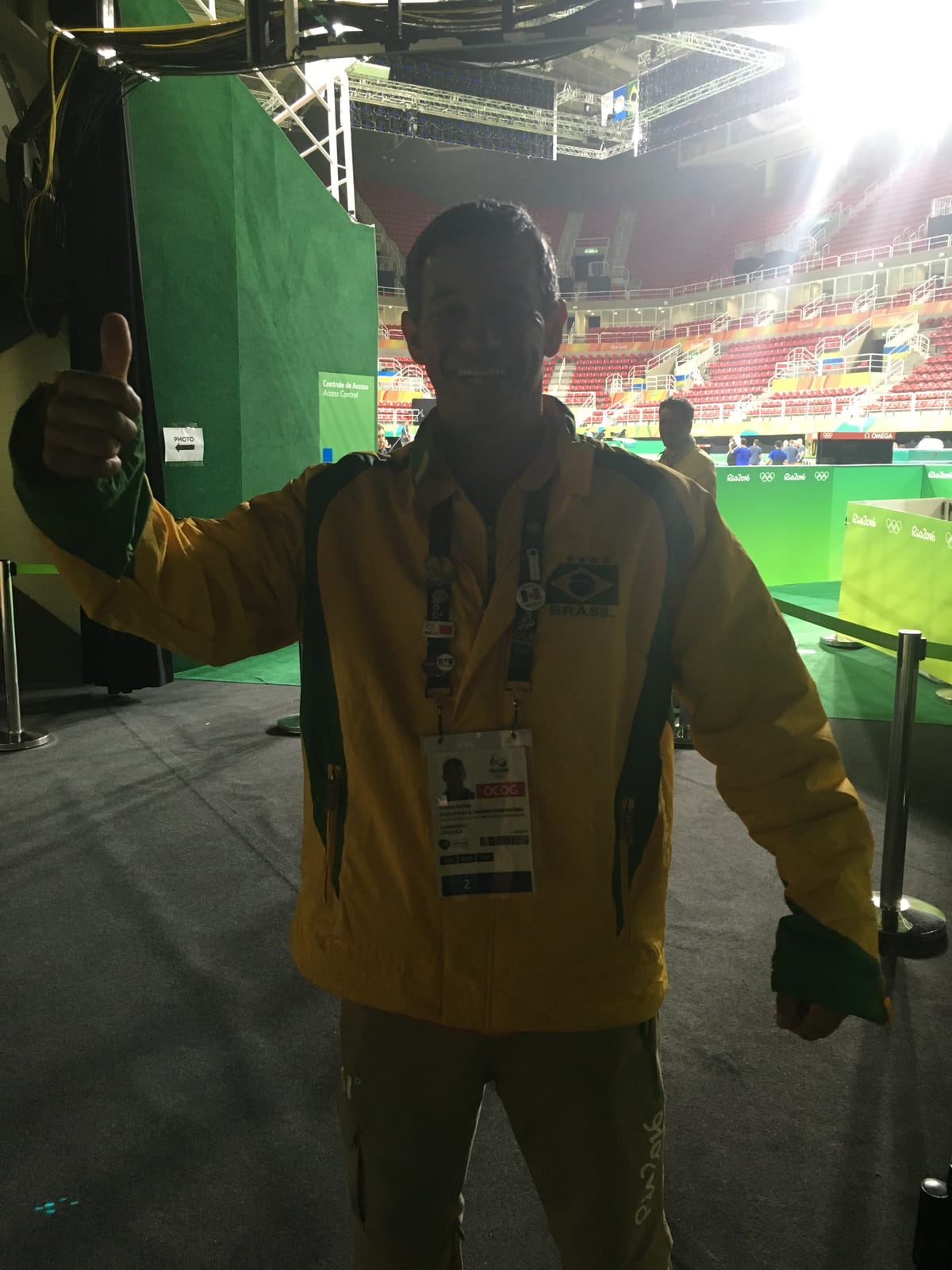A Rio 2016 volunteer in the yellow and green Brasil jacket giving a thumbs up in the empty gymnastics arena.