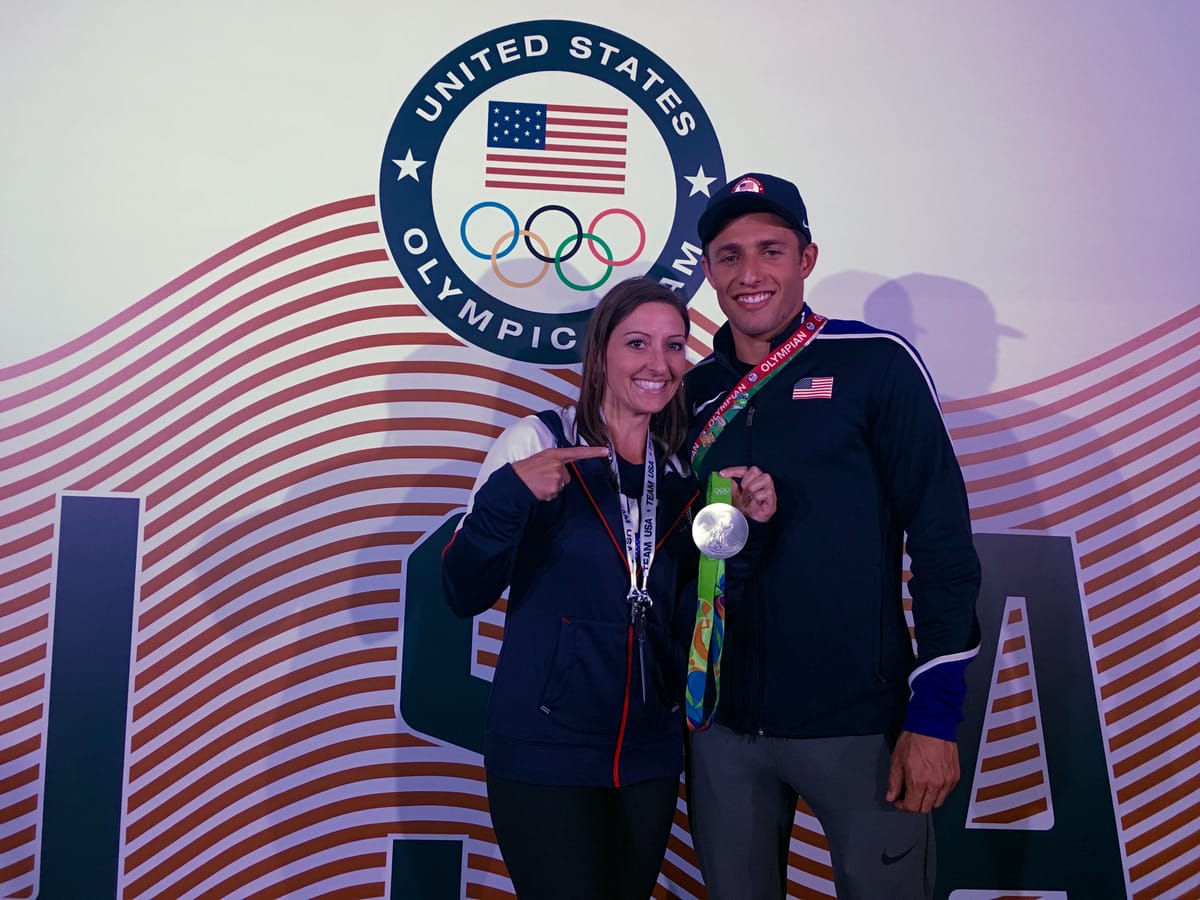 Gina Pongetti Angeletti pointing at the silver medal a US Olympian is holding up, both in Team USA kit in front of a US Olympic Team backdrop.