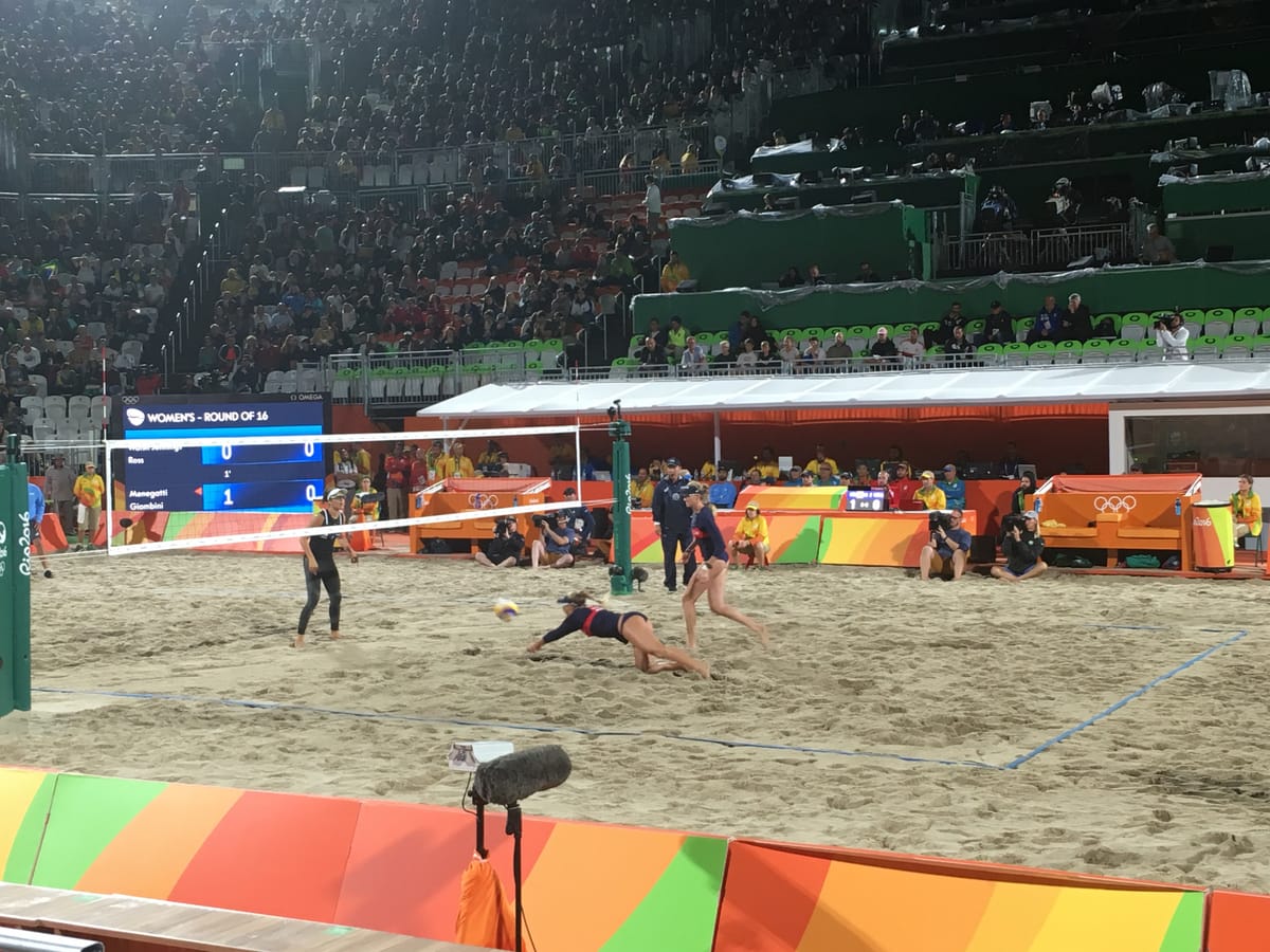 A women's beach volleyball round of sixteen match in the Rio arena, one player diving full length across the sand for the ball.