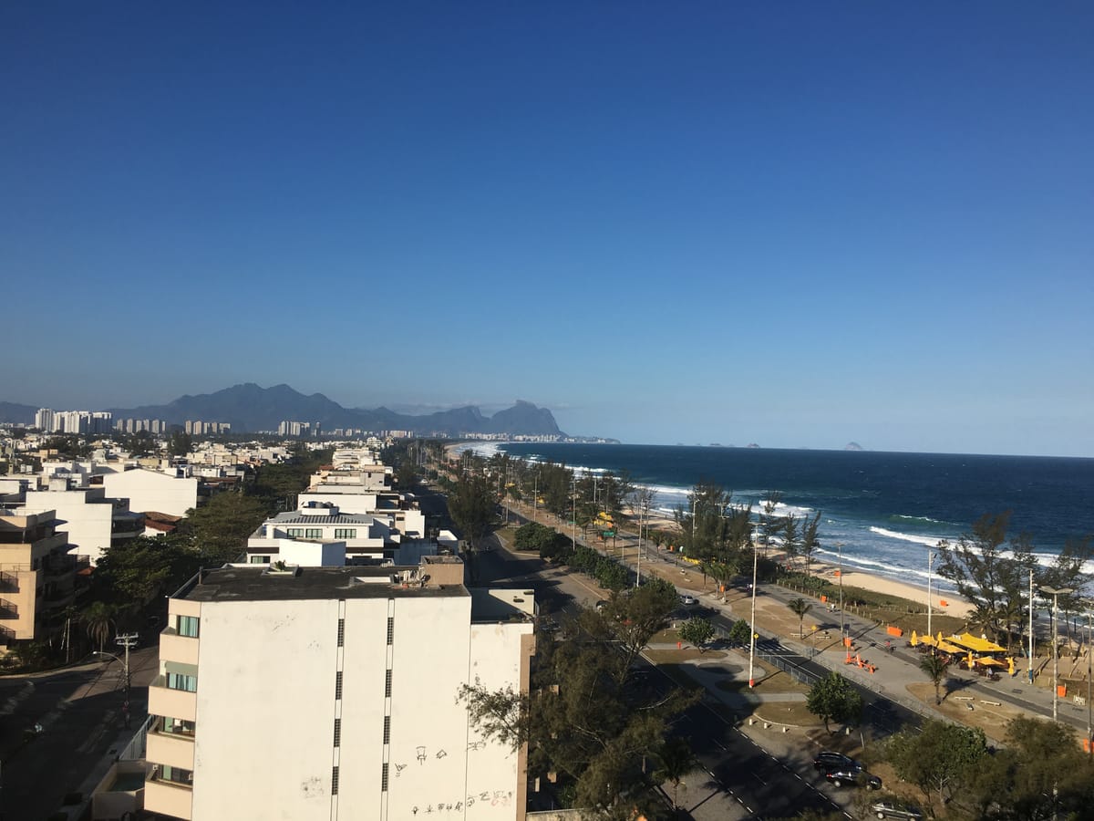 A high view along a Rio beachfront: apartment blocks, a wide highway, palm trees, and mountains in the far distance.