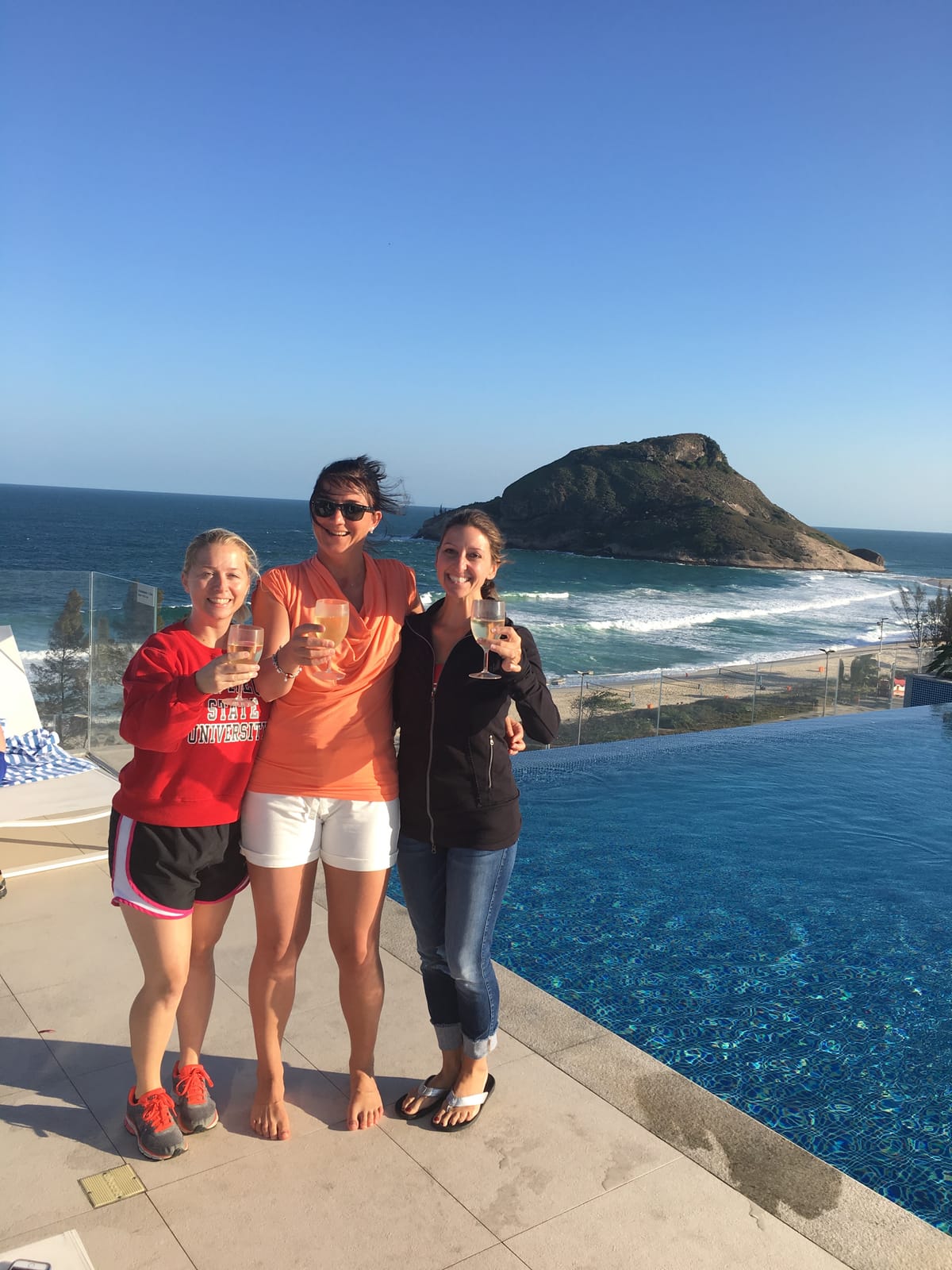 Three women raising glasses on a hotel terrace above an infinity pool, the ocean and a rocky headland behind them.