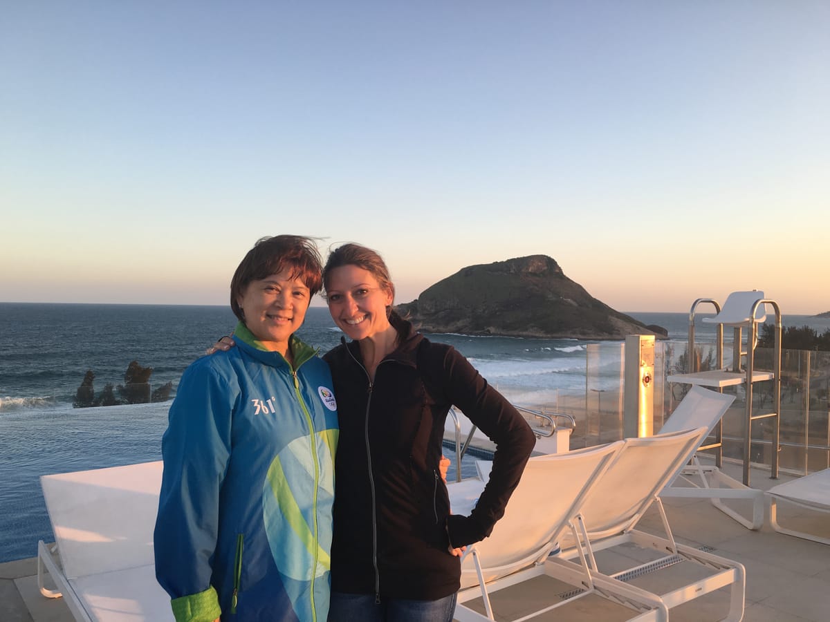 Gina Pongetti Angeletti with a woman in a blue Rio 2016 official's jacket on a terrace at sunset, the sea and a headland behind them.
