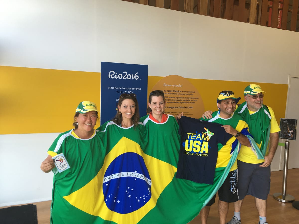 Five people at the Rio 2016 megastore wrapped in Brazilian flags and wearing Brasil caps, holding up a navy Team USA Rio de Janeiro t-shirt between them.