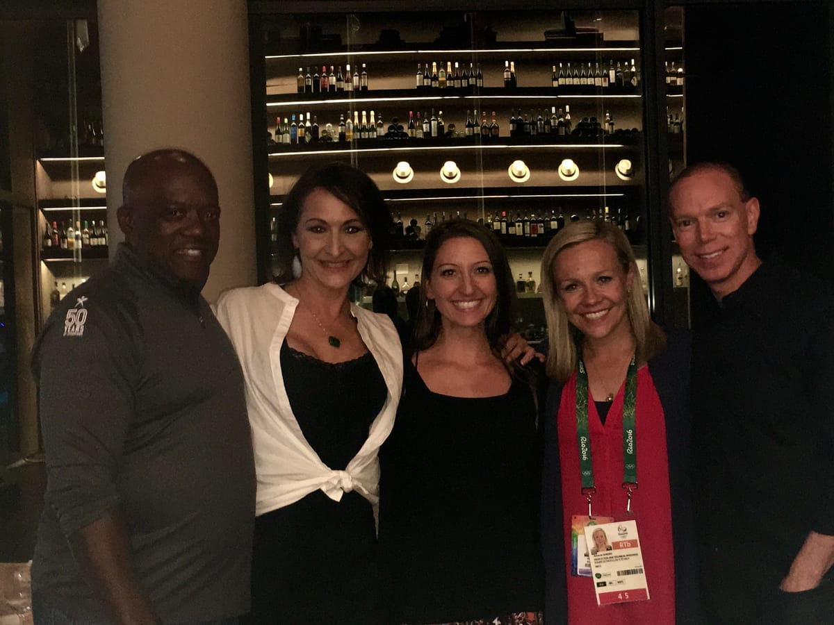 Five colleagues posed in front of a lit wine wall at a Rio bar, one still wearing a Games credential.