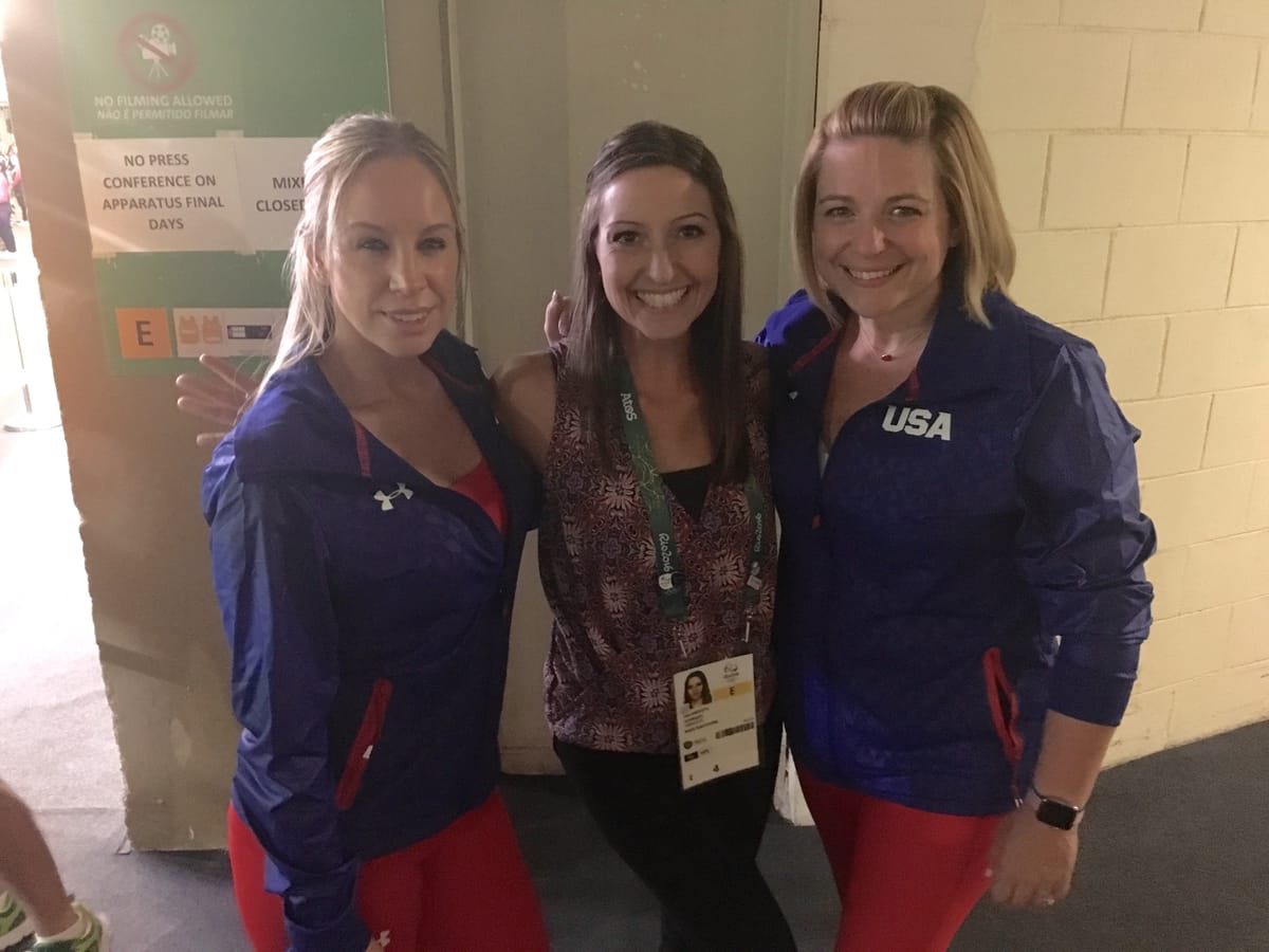 Gina Pongetti Angeletti between two US women's coaches in blue Team USA jackets in a back corridor of the arena, beneath a sign reading no press conference on apparatus final days.