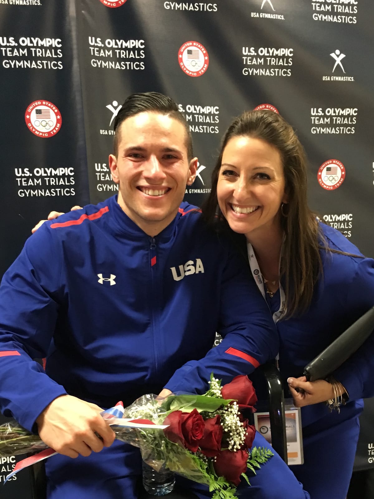 Gina Pongetti Angeletti alongside a US men's gymnast in a Team USA jacket holding a bouquet of red roses, at a US Olympic Team Trials backdrop.