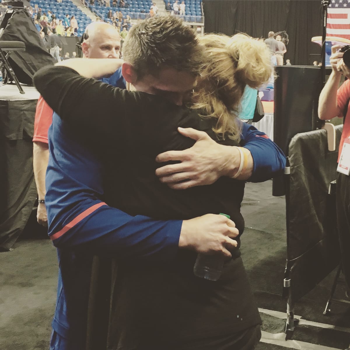 A man in a blue Team USA warm-up and a woman in black holding each other in a long hug on the competition floor.