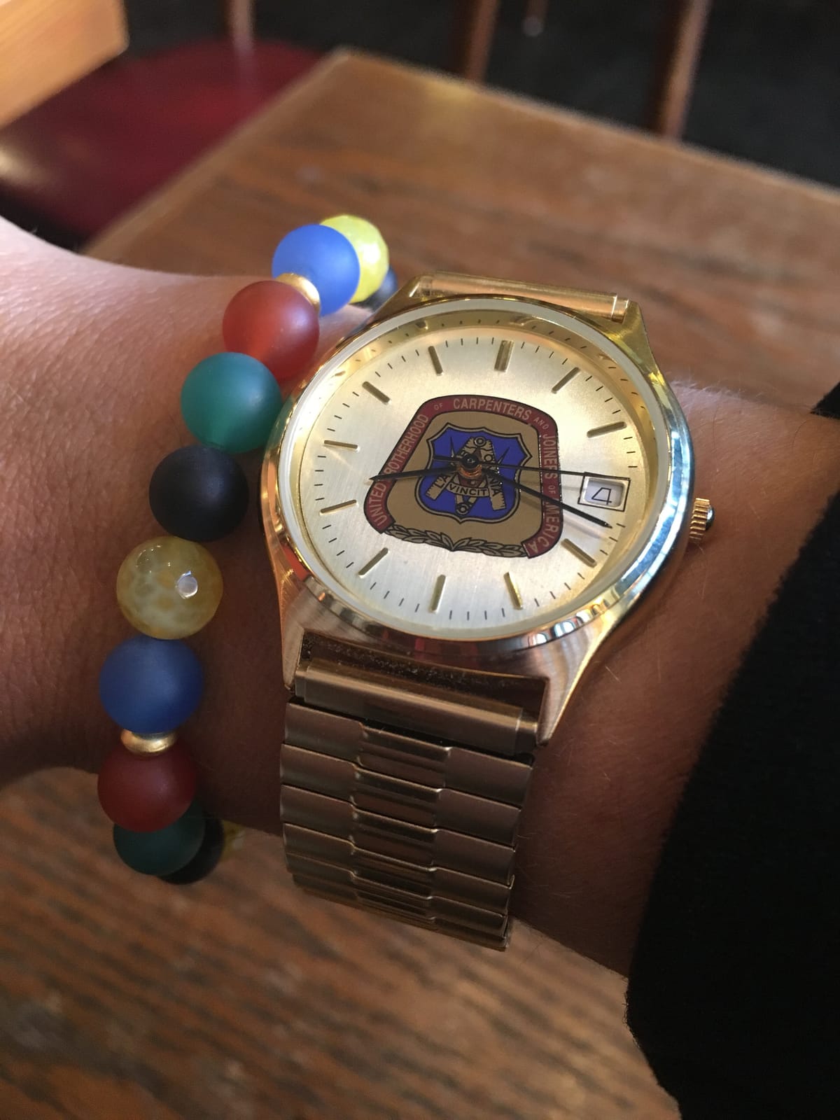 A gold United Brotherhood of Carpenters watch worn on a wrist, next to a bracelet strung with beads in the Olympic ring colours.