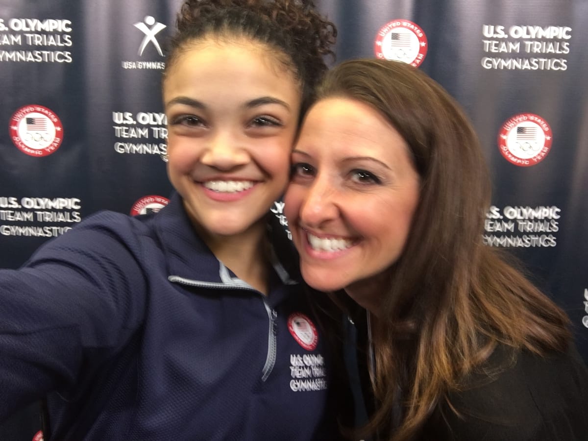 Gina Pongetti Angeletti taking a selfie with a young US gymnast in a Team USA jacket in front of a US Olympic Team Trials backdrop.