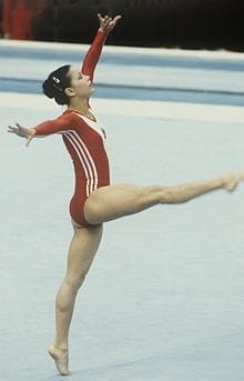 An archive photograph of Nellie Kim competing on floor exercise in a red and white leotard, one arm raised and one leg extended behind her.