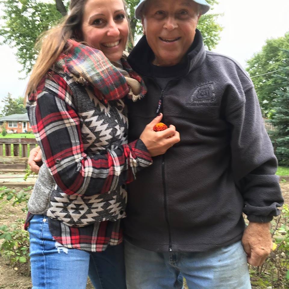 Gina Pongetti Angeletti with her father in a garden, her arm around him and an orange flower held against his chest.